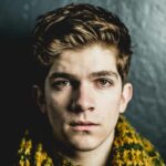 Close-up portrait of a young adult male with a serious expression against a dark textured wall.